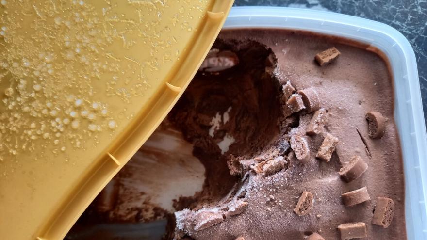 Eine geöffnete Schokoladeneispackung steht auf der Küchenarbeitsplatte. Auf der Deckelinnenseite und auf der Oberfläche des Speiseeises sind Eiskristalle zu sehen.